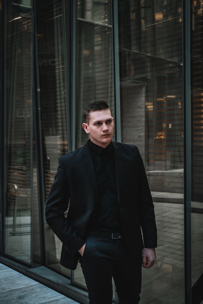 Young man in sleek dark suit stands confidently by a modern glass building facade.