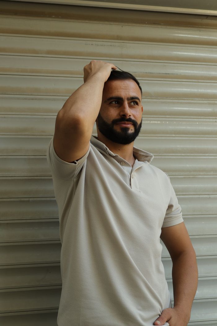 Portrait of a bearded man in a white polo shirt posing against a beige wall.