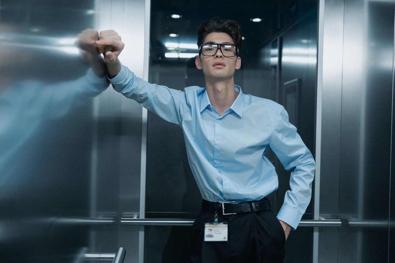 Portrait of an Asian man in blue long sleeves with eyeglasses, leaning in an elevator.