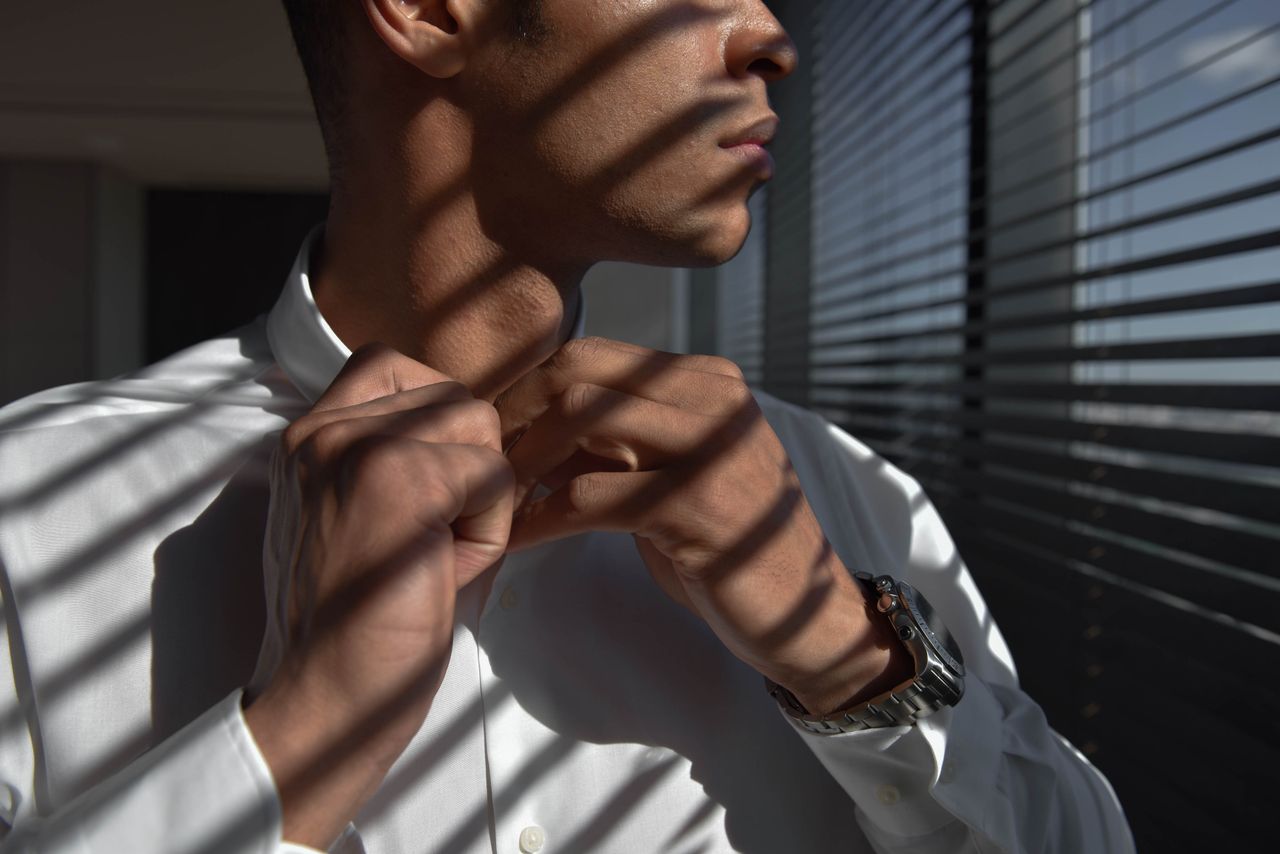 Close-up of a man adjusting his shirt with sunlit shadows creating a patterned effect indoors.